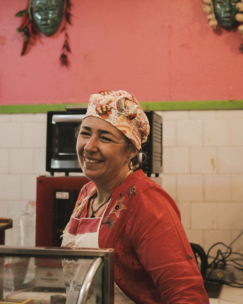 Cozinheira sorrindo atrás do balcão de uma cozinha pequena.